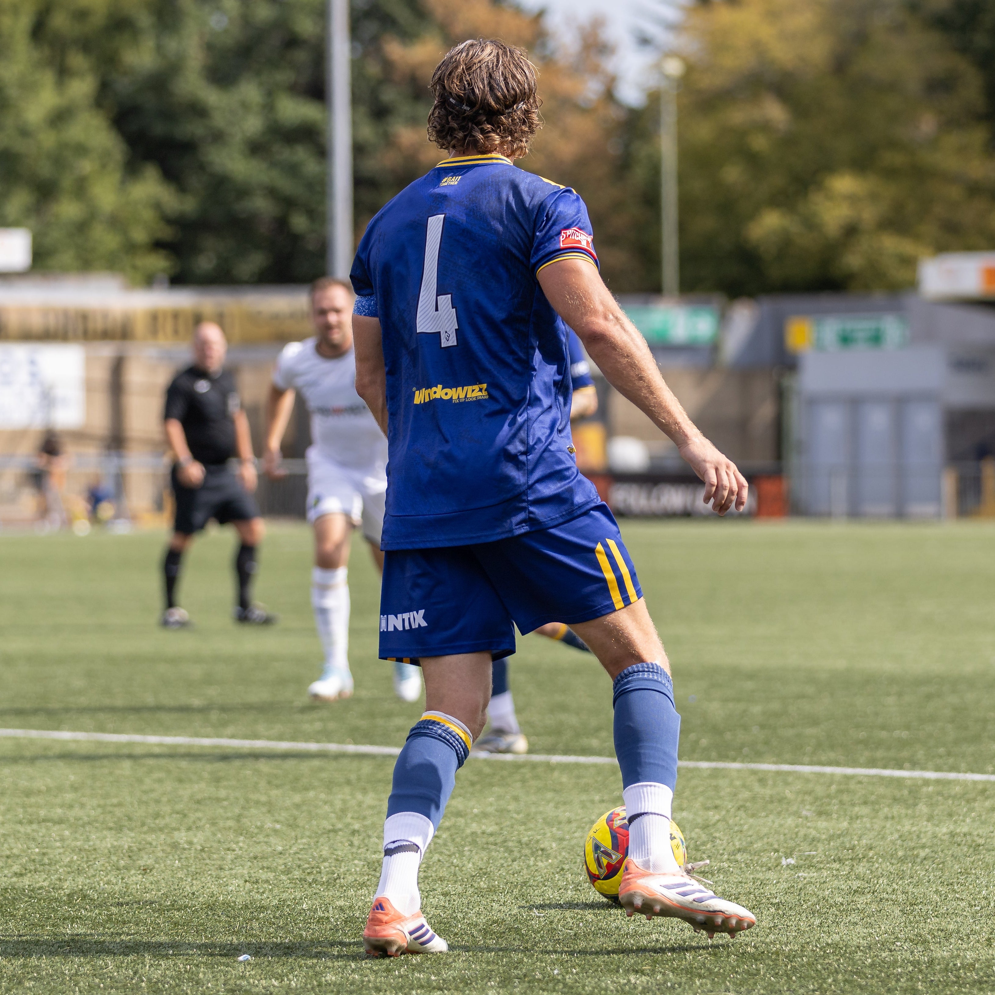 A soccer player wearing a blue jersey with the number 4 and matching blue shorts stands on a green turf field, facing away from the camera. Another player in a white kit is visible in the background near the center, with trees and stadium structures blurred in the distance.