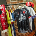 A black athletic jersey with a white splatter pattern and 'Ba11ers' text is displayed on a clothing rack alongside other sports jerseys, a red scarf, and a black cap in a rustic indoor setting with a brick wall background.
